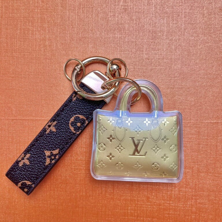 A keychain featuring a clear acrylic mini shopping bag charm with embossed gold-tone pattern and a dark navy monogram leather strap, attached to gold-tone rings on an orange textured background.